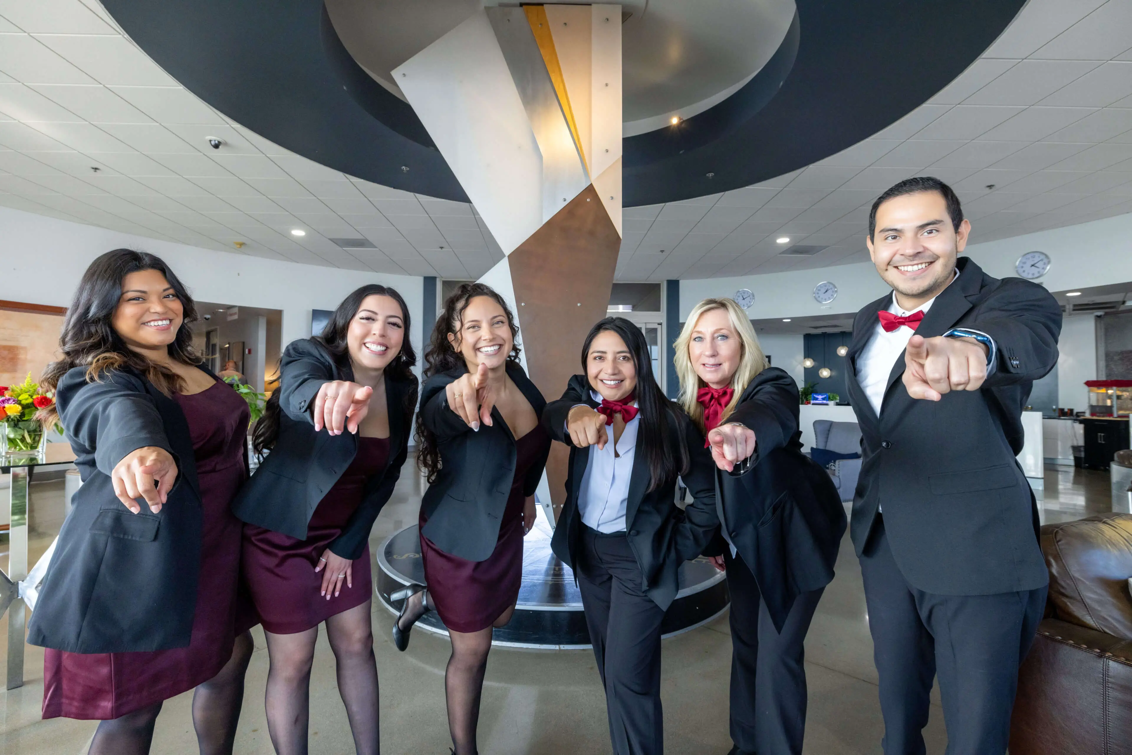 A group of six professionals in formal attire smiling and pointing forward.
